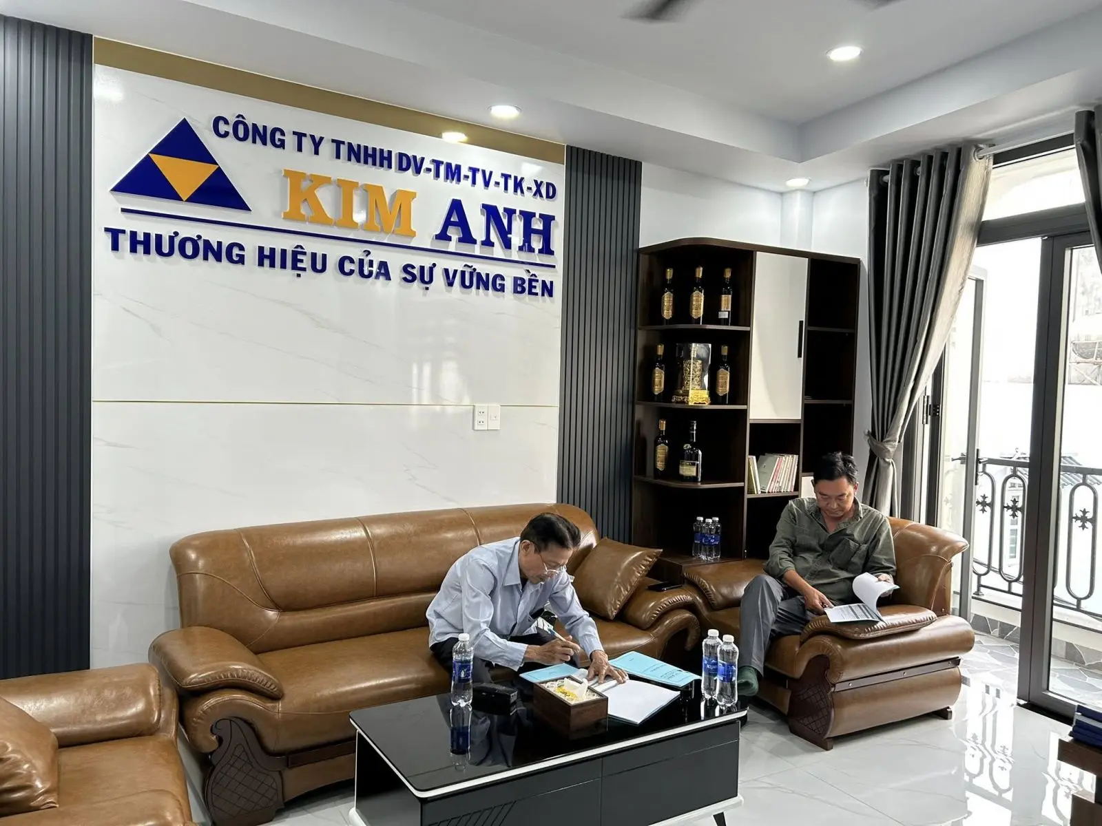 Một gia chủ đang ngồi đối diện với giám đốc nhà thầu tại văn phòng, trên bàn là bản vẽ thiết kế và bảng báo giá chi tiết, không khí buổi làm việc nghiêm túc, chuyên nghiệp